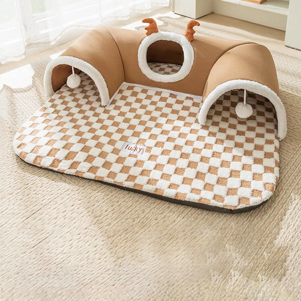 Camel cat bed with a curved tunnel, two hanging pompoms and a checked cream mat, set on a beige rug