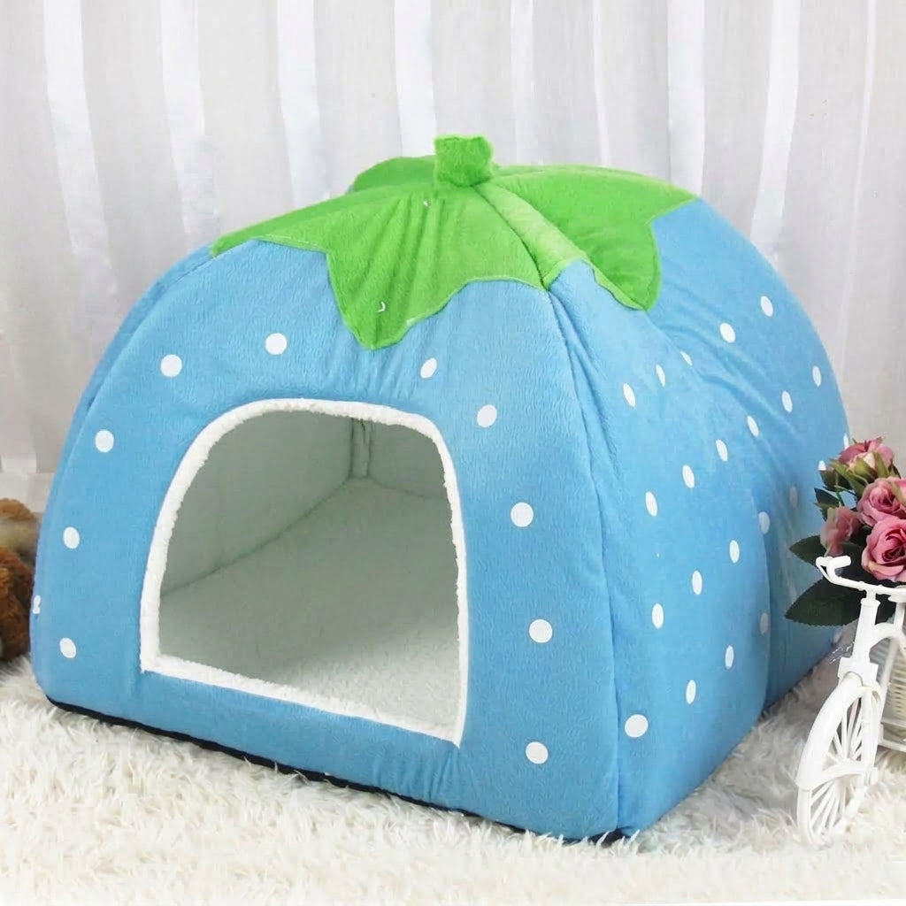 Blue strawberry cat cave bed with white dots and a green leaf top, on a white fluffy rug beside pink flowers