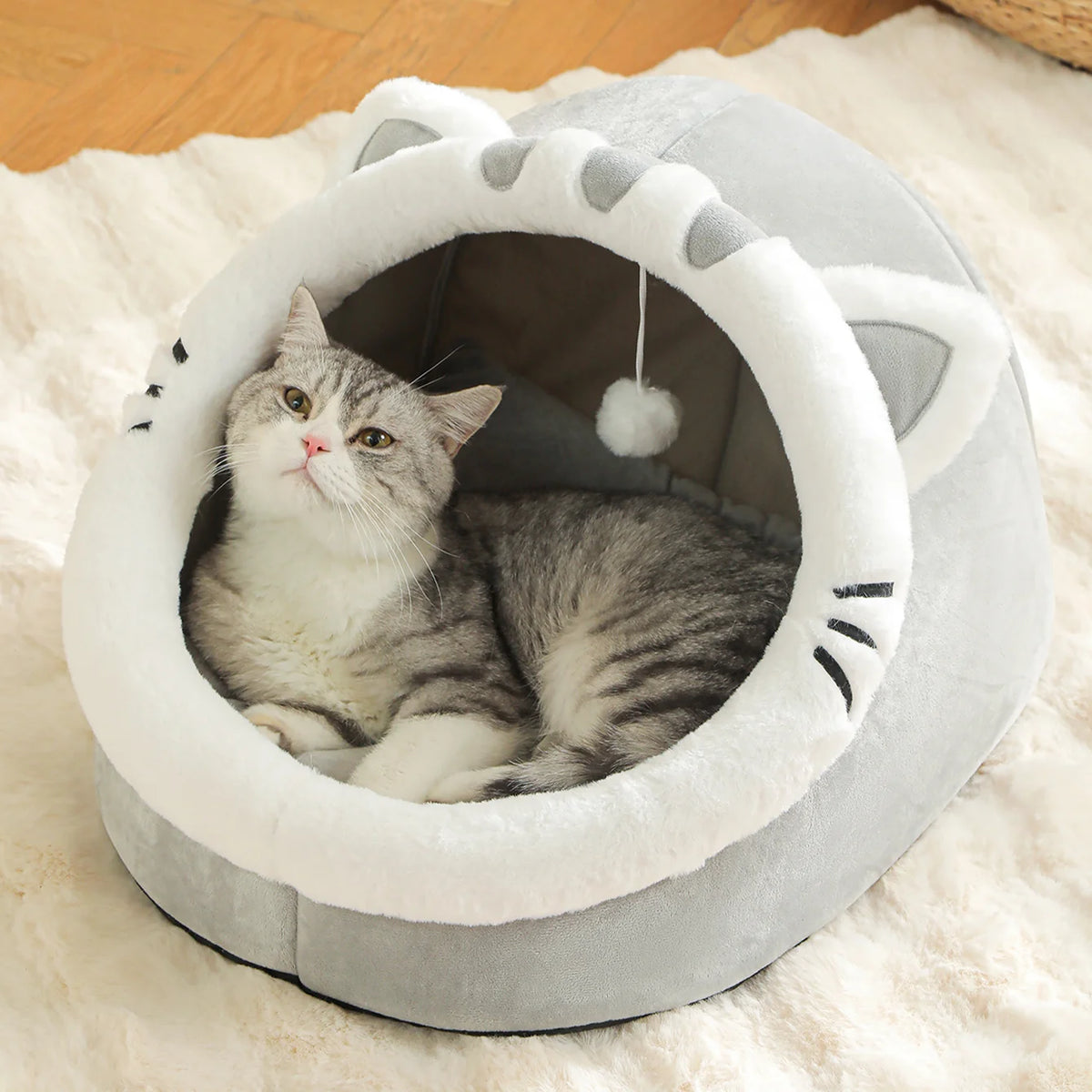 Grey and white tabby cat lying inside a grey cat face basket bed with ears and whiskers on a cream fluffy rug