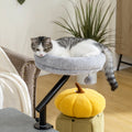 Tabby and white cat lying in a grey plush desk clamp cat bed fixed to the edge of a table beside an armchair