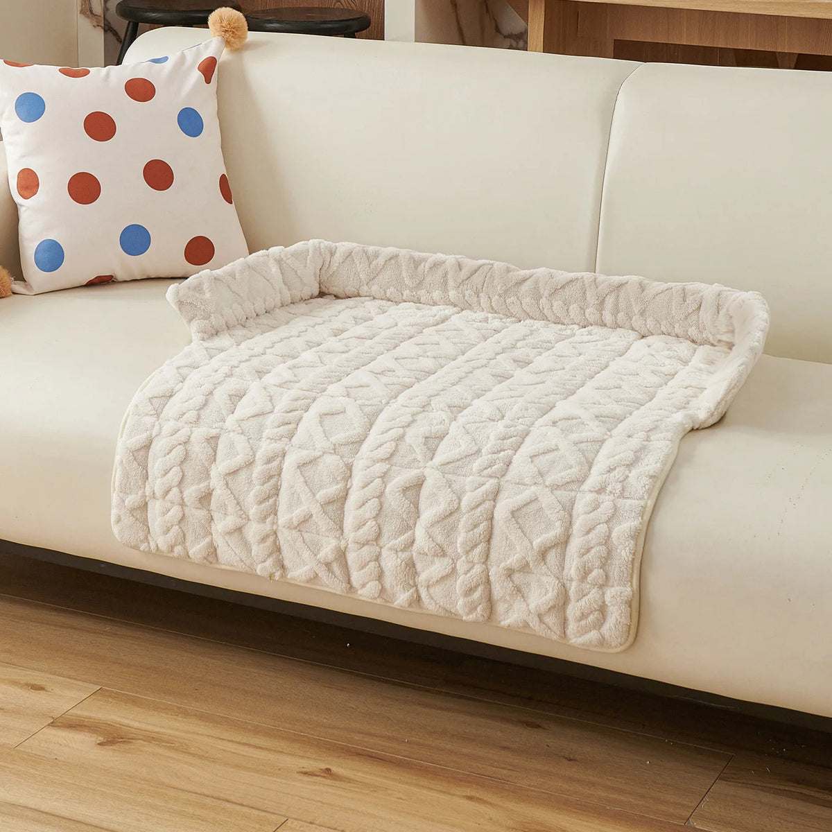 Cream cable knit sofa pad with a plush bolster, laid over the seat of a cream sofa beside a spotted cushion