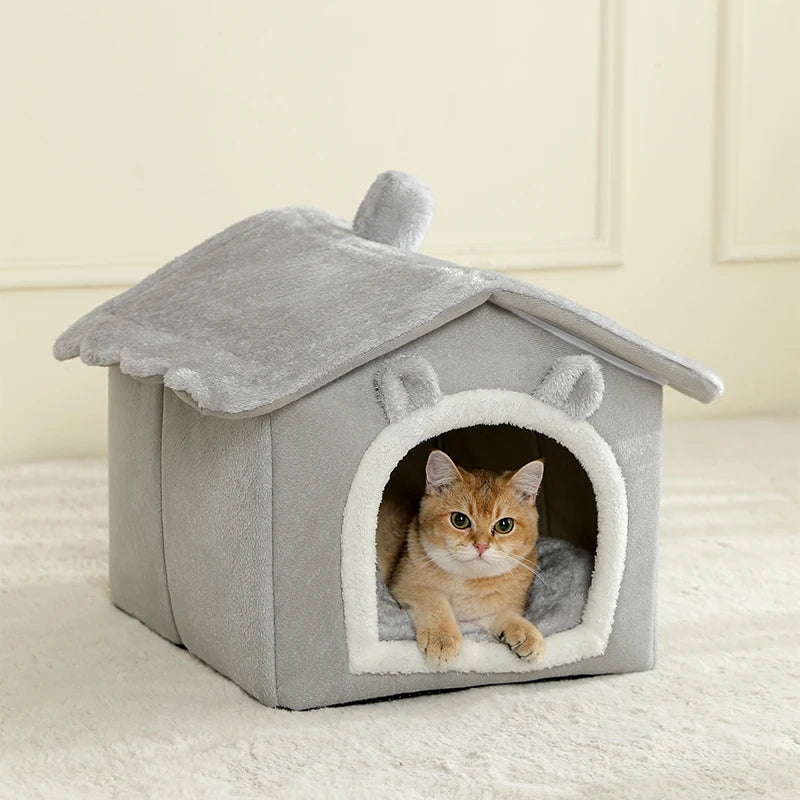 Ginger cat peeking out of the arched doorway of a grey house-shaped hooded pet bed on a cream rug