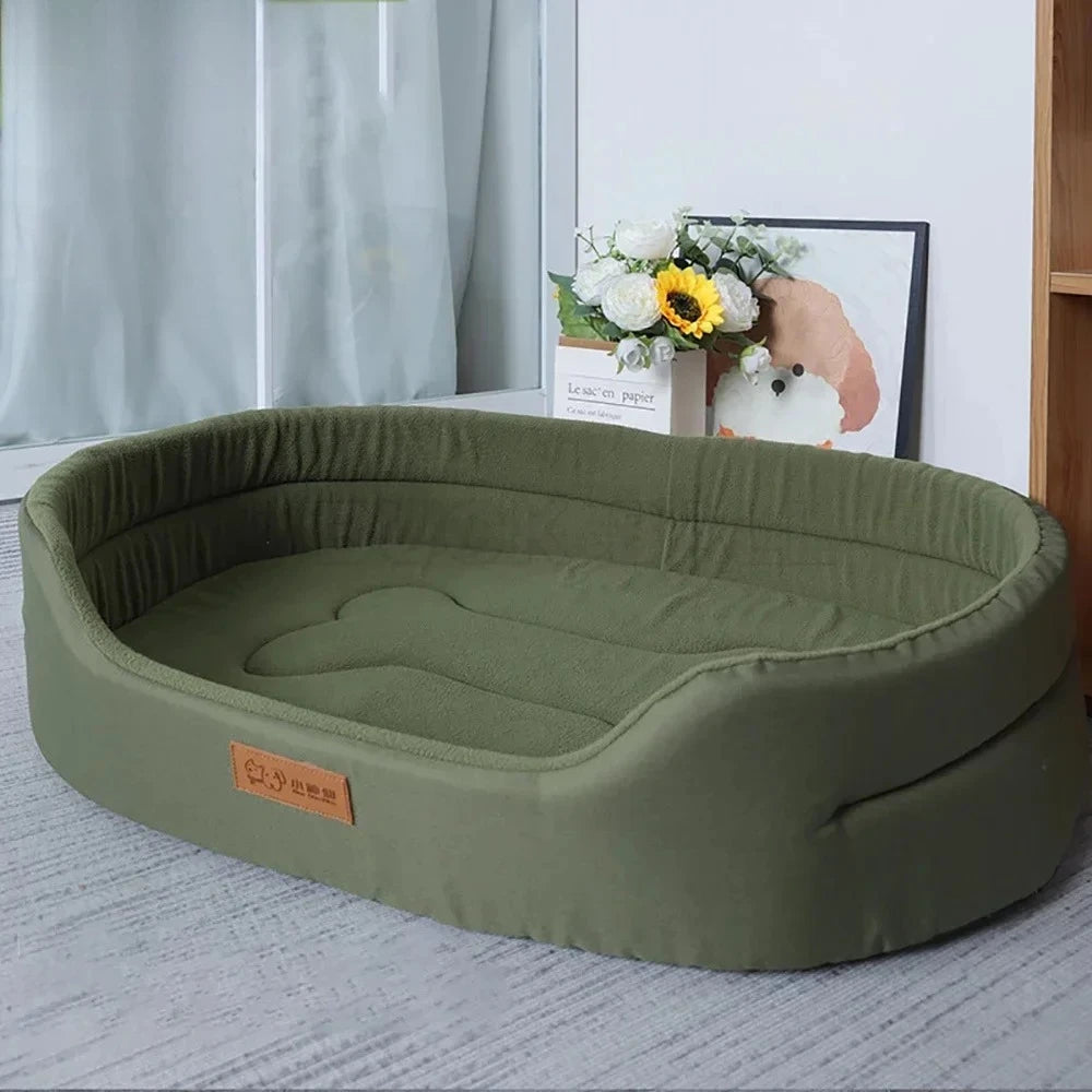 Olive green oval dog bed with high sides on a grey rug, flowers and a framed dog picture behind it