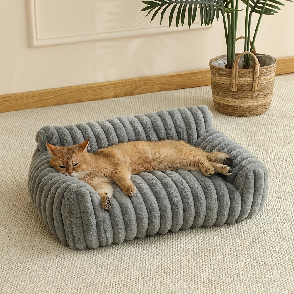Ginger cat stretched out asleep on a grey ribbed plush cat couch bed on a cream rug beside a potted plant
