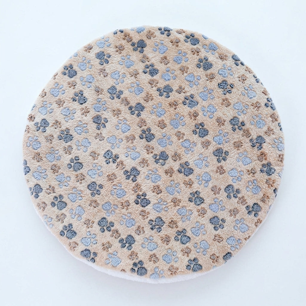 Round cat mat in beige plush printed with grey, blue and brown paw prints, on a pale blue-grey background