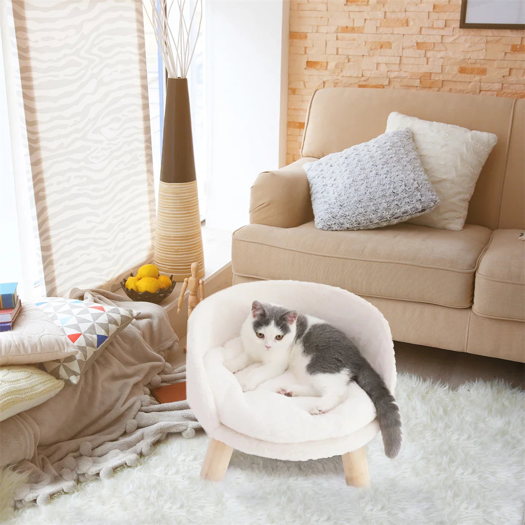 Grey and white cat lying in a cream plush round sofa bed on wooden legs, on a white rug beside a beige sofa