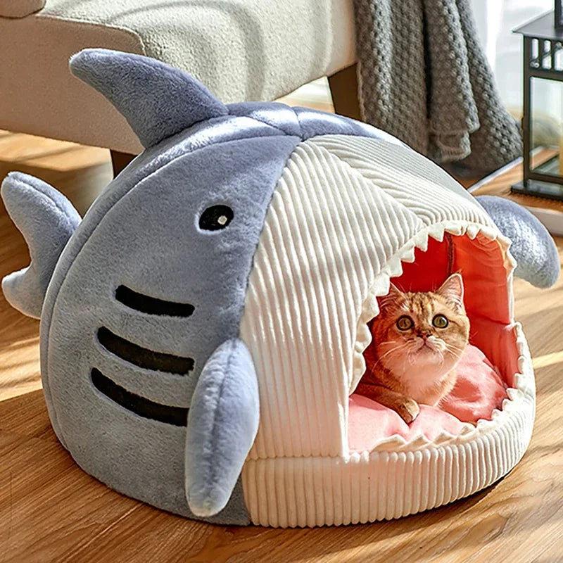 Blue shark cat bed cave on a wooden floor, a ginger kitten lying on the pink cushion inside its open mouth
