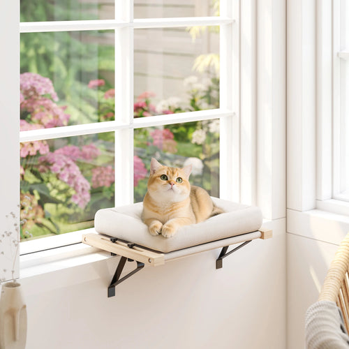 Ginger cat sitting on a cream windowsill cat bed fixed to a white window ledge, a flowering garden outside